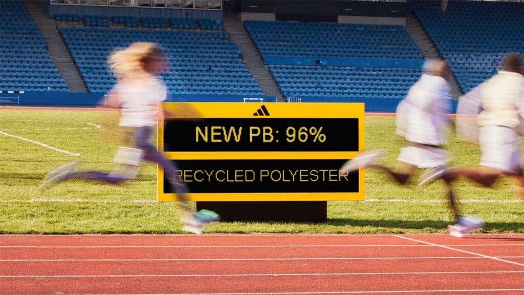 Cuatro corredores pasan corriendo junto a un cartel digital en el campo de atletismo que indica "NUEVA PB: 96% POLIÉSTER RECICLADO", con los asientos azules vacíos del estadio al fondo.