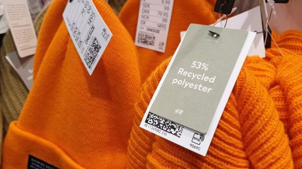 Primer plano de gorros de punto naranja con etiquetas que indican que están fabricados con un 53% de poliéster reciclado, promoviendo la circularidad en la moda.