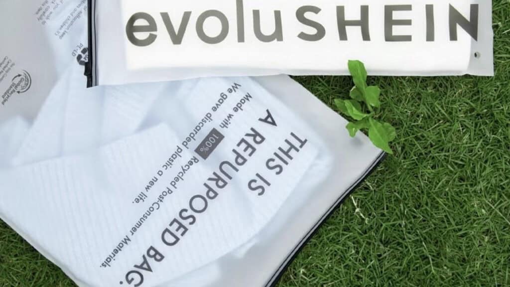 Una bolsa reutilizable con la etiqueta "ESTA ES UNA BOLSA REPURSADA" y un artículo doblado con la etiqueta "evoluSHEIN", fabricado con poliéster reciclado, descansan sobre la hierba verde junto a una pequeña planta.