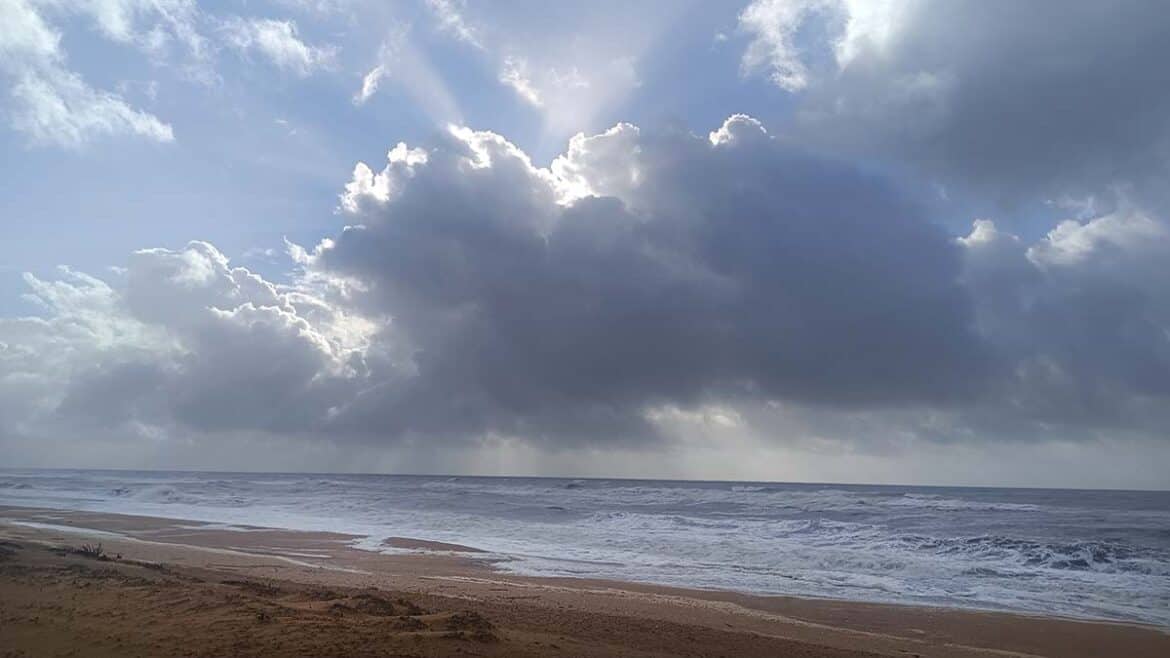 Las nubes bloquean parcialmente el sol sobre una playa de arena mientras las olas se acercan a la orilla, creando un cielo dramático con rayos de luz que se abren paso.