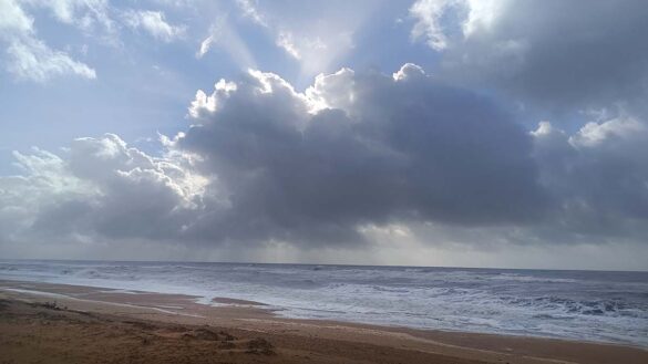 Las nubes bloquean parcialmente el sol sobre una playa de arena mientras las olas se acercan a la orilla, creando un cielo dramático con rayos de luz que se abren paso.