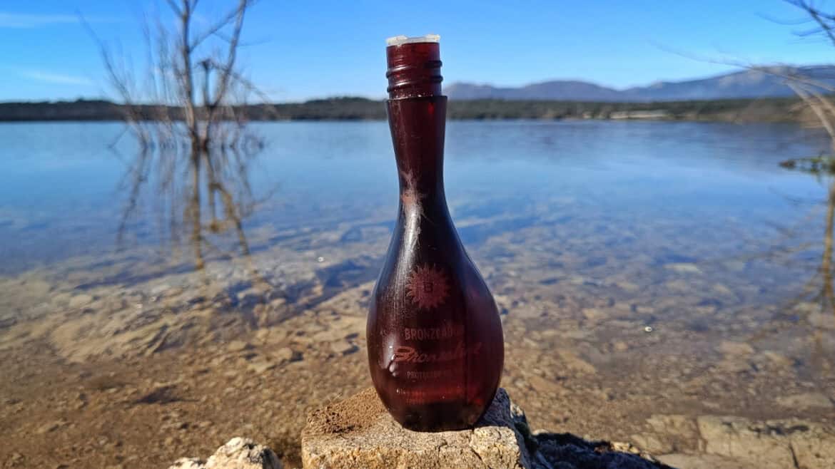 Una botella de Brozaline de vidrio marrón con letras descoloridas se alza erguida sobre una roca a la orilla de un lago claro y tranquilo, evocando el ambiente apacible de los anos 70, con las montañas elevándose al fondo.