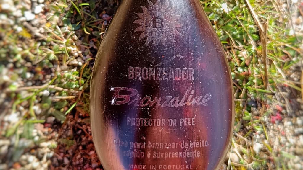 Primer plano de un frasco de aceite bronceador protector de la piel Bronaline, entre hierba y pequeñas plantas. La etiqueta, con la marca "Borrador automático", destaca su origen: fabricado en Portugal.