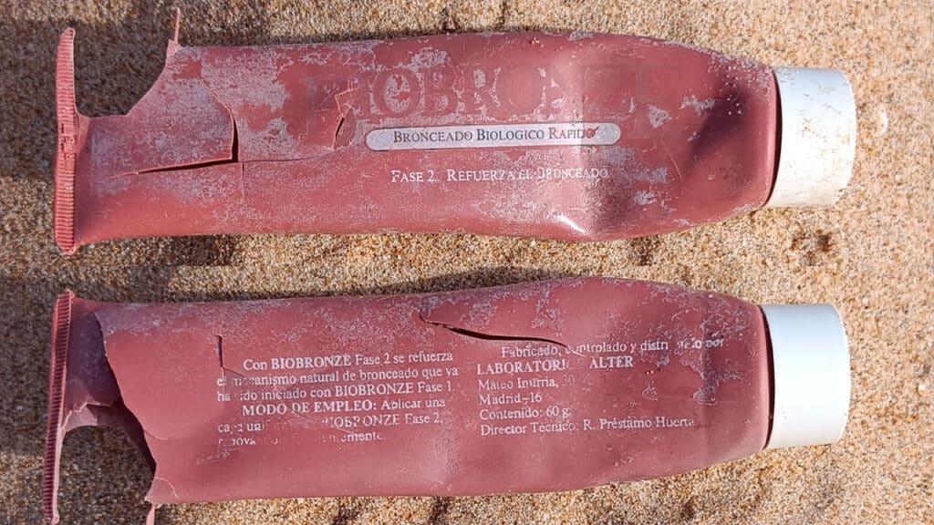 Dos tubos rojos usados de crema solar o bronceadora, etiquetados en español, yacen en la arena con la superficie desgastada y el texto descolorido, casi como si hubieran sido tocados por un borrador automático natural.
