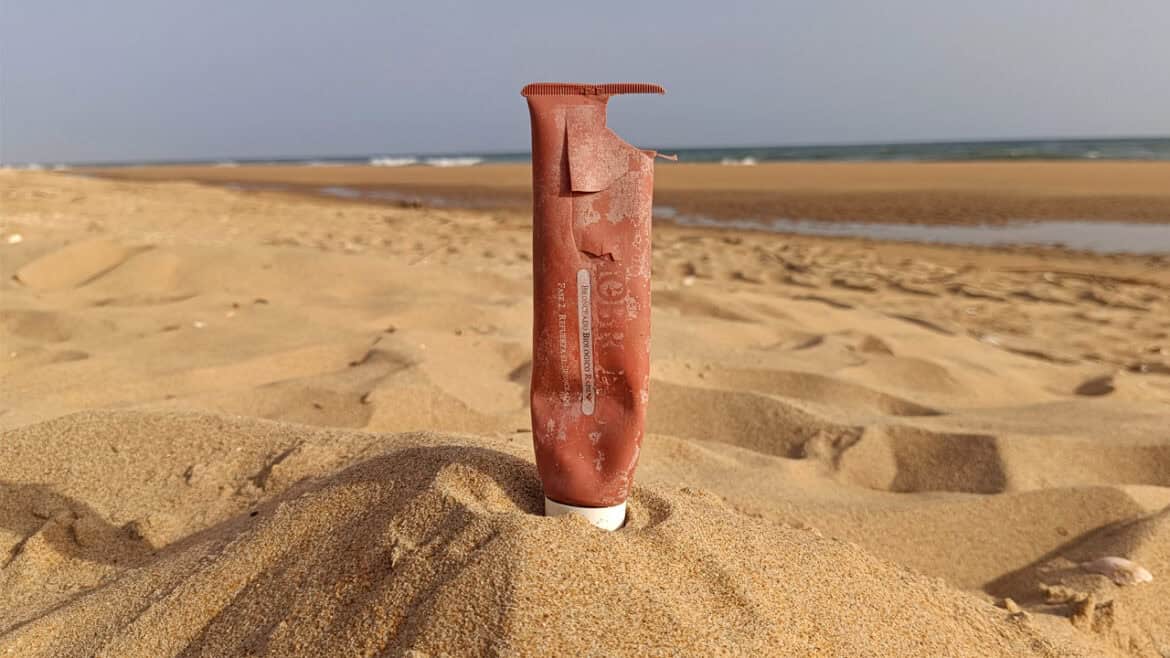 Un tubo de plástico dañado y vacío -como un borrador automático- parcialmente enterrado en posición vertical en la arena de la playa, con el mar y el cielo azul de fondo.
