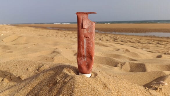 Un tubo de plástico dañado y vacío -como un borrador automático- parcialmente enterrado en posición vertical en la arena de la playa, con el mar y el cielo azul de fondo.