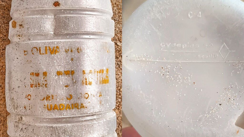 Primer plano de una botella de aceite de oliva de los años 70 encontrada en la playa, transparente y desgastada, con un texto amarillo descolorido en el lateral y marcas en relieve en la parte inferior, ambas parcialmente ocultas por arañazos y arena.