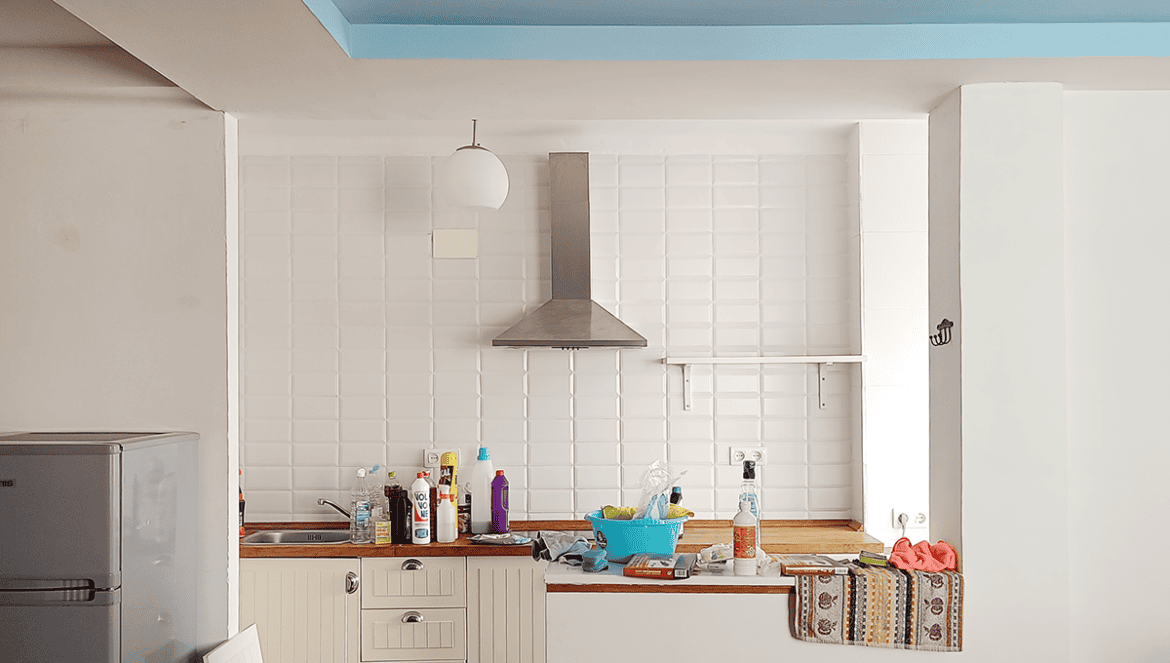 Una pequeña cocina con artículos de limpieza sobre la encimera, una campana extractora de acero inoxidable, un frigorífico y paredes de azulejos blancos: cada rincón parece contar su propia historia de la vida cotidiana.