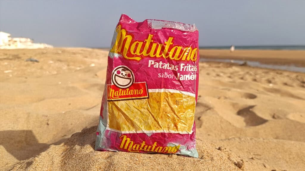Una bolsa arrugada de patatas fritas con sabor a jamón Patatas Matutano se yergue sobre la arena de la playa, evocando el espíritu despreocupado de los años 80 con el mar visible al fondo.