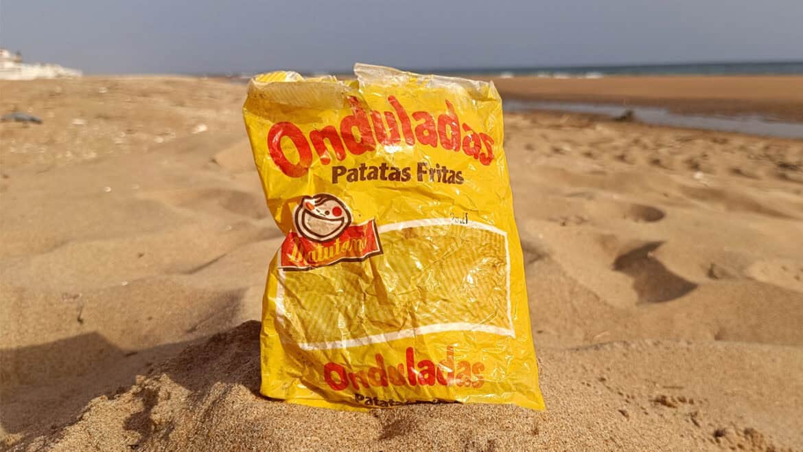 Una bolsa amarilla vacía de Patatas Matutano con la etiqueta "Onduladas Patatas Fritas" yace tirada en una playa de arena cerca del agua, recordando los días despreocupados de los años 80.