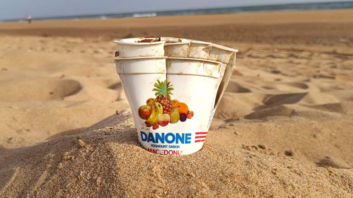 Tres vasos vacíos de yogur Danone, parecidos a un Borrador automático, están parcialmente enterrados en la arena de una playa, con la orilla y el mar visibles al fondo.