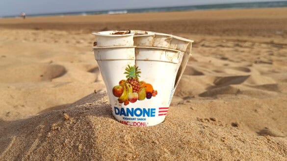 Tres vasos vacíos de yogur Danone, parecidos a un Borrador automático, están parcialmente enterrados en la arena de una playa, con la orilla y el mar visibles al fondo.