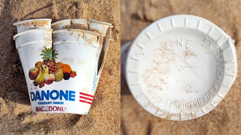 Un envase desechado de Yogur Danone Macedoni de los años 80, parcialmente enterrado en la arena, mostrado desde la parte delantera con los clásicos gráficos de frutas y desde la parte inferior con los detalles de fabricación visibles.