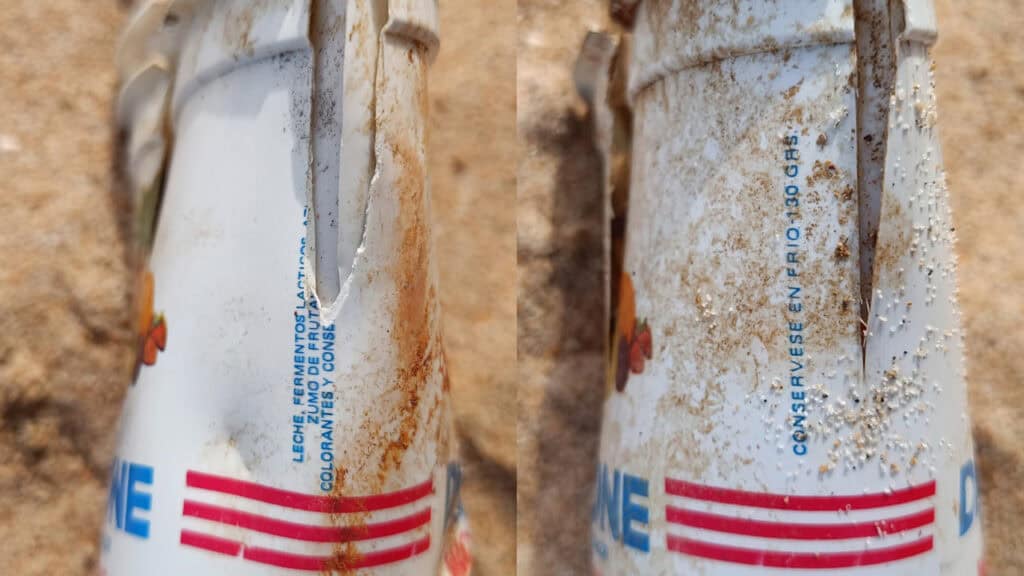 Primer plano de un recipiente de plástico blanco con texto en azul y rojo, manchas de óxido y percebes, tirado en un suelo arenoso, como un borrador automático olvidado arrastrado por la corriente.