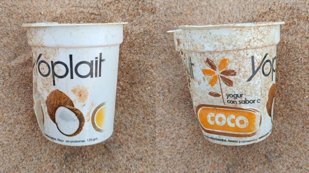 Dos envases vacíos y desgastados de yogur Yoplait con etiquetado de sabor a coco, probablemente desechados como borrador automático, están clavados en la arena, mostrando suciedad y desgaste.