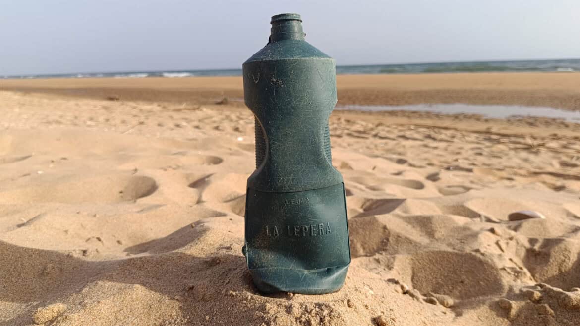 Una botella de plástico desgastada, como un borrador automático olvidado, yace parcialmente enterrada en la arena de una playa, con el océano y el cielo extendiéndose apaciblemente al fondo.