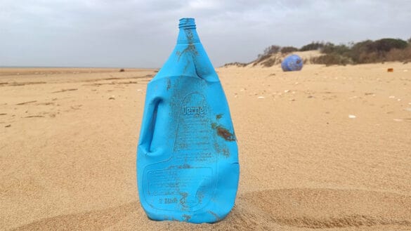 Un contenedor de plástico azul, con la inscripción "Borrador automático", está parcialmente enterrado en la arena de una playa desierta con algo de vegetación al fondo.