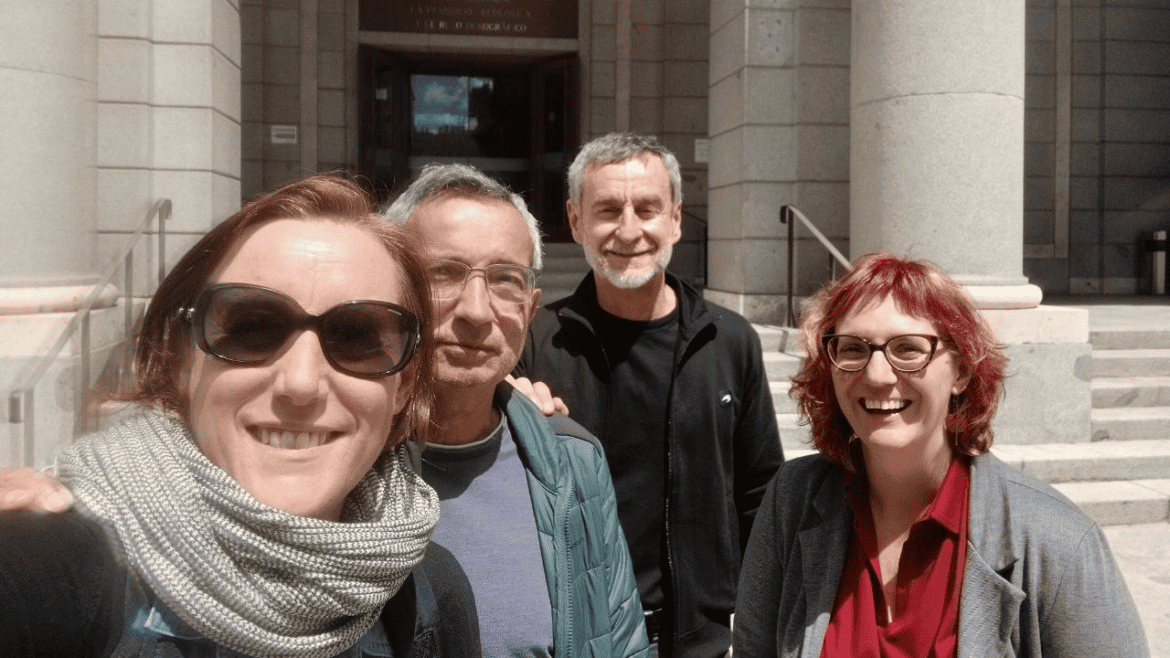 Cuatro adultos posan y sonríen para un selfie de grupo en el exterior de un edificio de piedra con grandes columnas y escaleras, capturando el momento en un borrador automático.