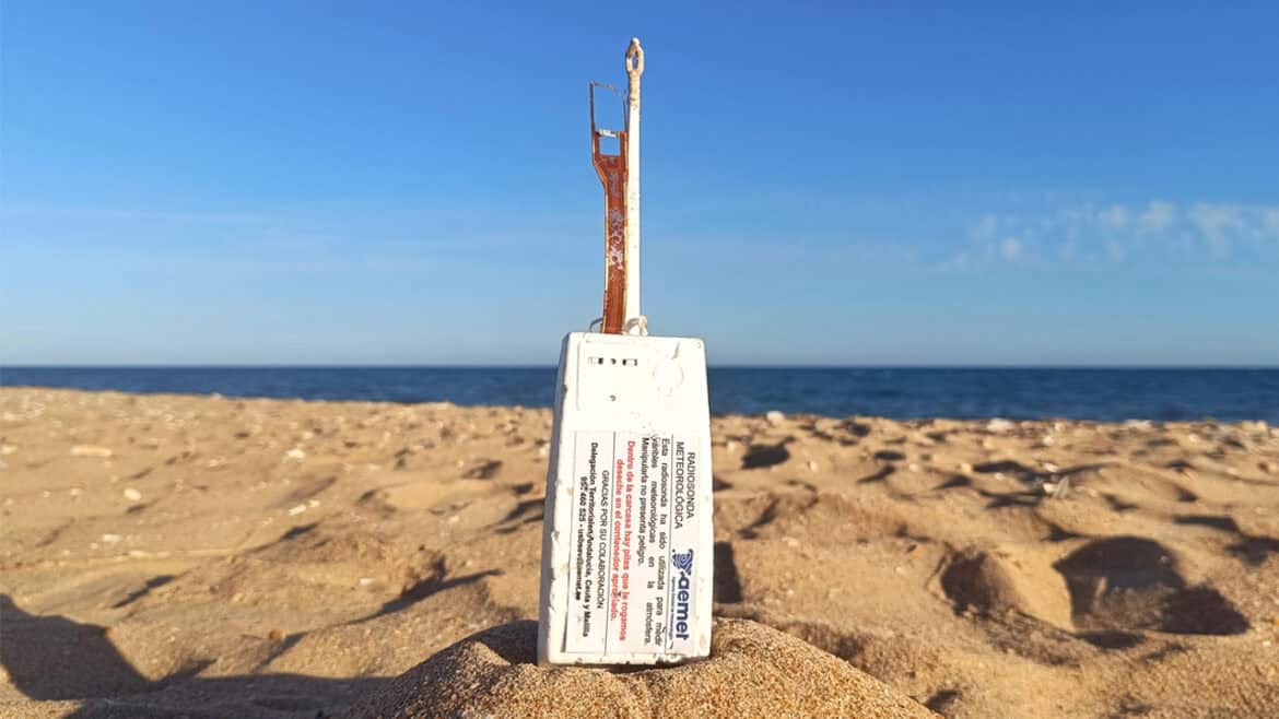 Un aparato de plástico desechado y desgastado, con piezas metálicas expuestas, yace parcialmente enterrado en la arena de la playa, parecido a un borrador automático olvidado, mientras el océano y el cielo se extienden al fondo.