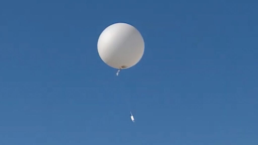 Un globo meteorológico blanco flota en un cielo azul despejado, con un pequeño paquete de instrumentos borrador automático sujeto por una cuerda por debajo.