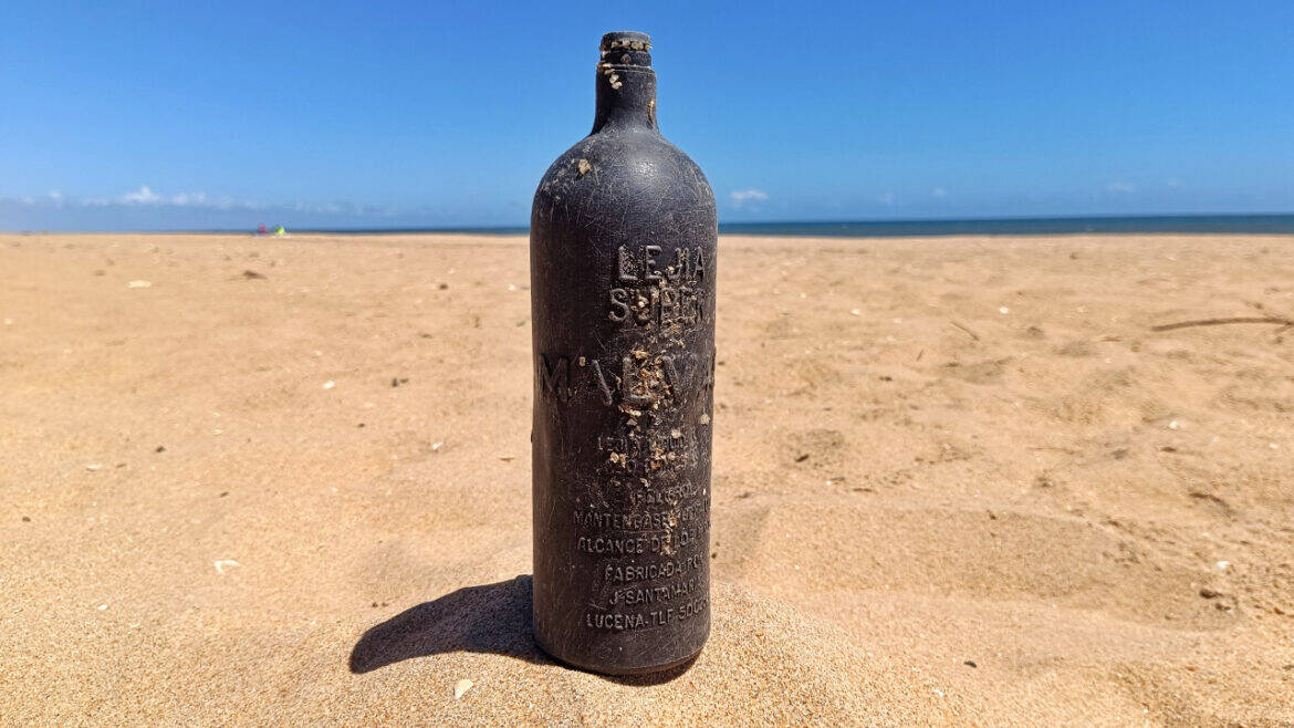 Una botella de cristal oscuro y desgastado se yergue sobre la arena de una playa desierta, con el océano y el cielo azul como telón de fondo: una escena que recuerda a una cámara automática captando un momento fugaz.