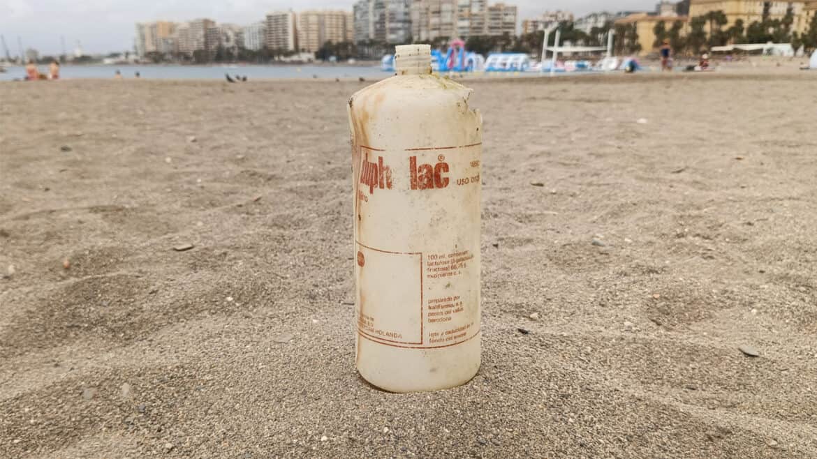 Una botella de plástico grande y desgastada, que se asemeja a un borrador automático, se yergue en la arena de una playa, con edificios de la ciudad y el mar visibles al fondo.