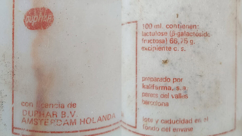 Primer plano de la etiqueta de un medicamento español en la que se muestran los ingredientes, los datos del fabricante y la información de uso, con parte del texto descolorido, como si se hubiera borrado con un borrador automático, y con marcas de suciedad visibles en la superficie.