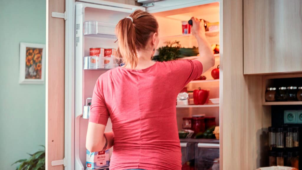 Una mujer con una camiseta roja está de pie delante de una nevera abierta, con un cartón de leche en la mano y tratando de alcanzar el pescado que hay guardado en envases de plástico en el estante superior.
