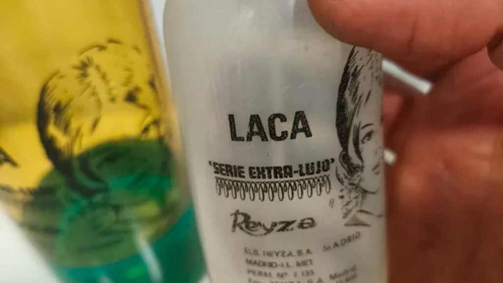 Primer plano de una mano que sostiene una botella blanca de estilo vintage con la etiqueta «Laca», que incluye texto en español y la ilustración del rostro de una mujer; al fondo se ve una botella amarilla y verde de Envase Pervisa de los años 80.