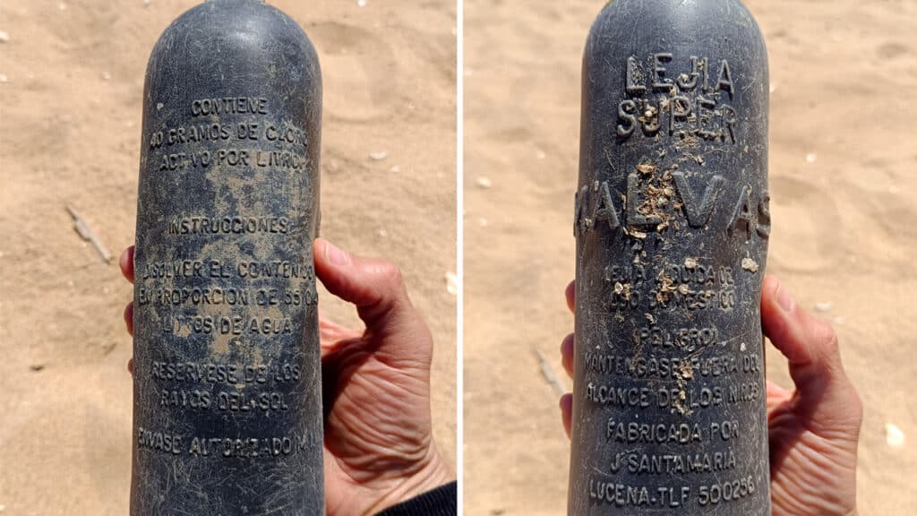 Una mano sostiene una botella vieja y desgastada de color gris, con un texto en español desdibujado y una etiqueta en la que apenas se lee «Borrador automático»; la botella aparece fotografiada desde dos ángulos en una playa de arena.