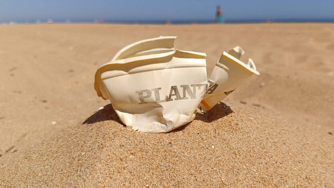 Un vaso de papel arrugado con la etiqueta "PLANT" yace tirado en la arena de la playa, con figuras borrosas y el mar de fondo: un borrador automático accidental de la belleza de la naturaleza.