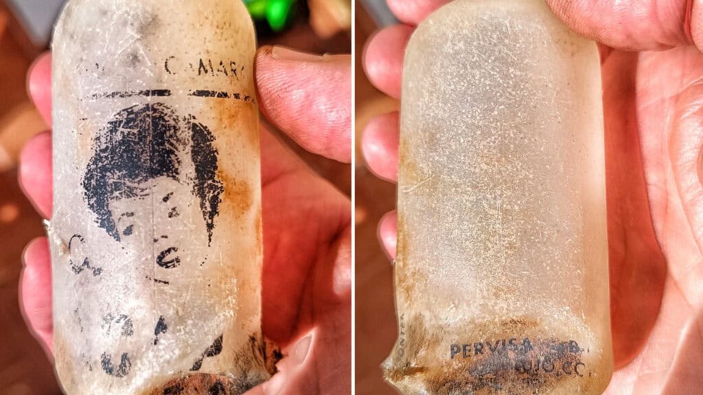 Una mano sostiene una vieja botella de cristal esmerilado con una ilustración descolorida del rostro de un niño y un texto parcial e ilegible. En el lado opuesto se aprecia más escritura descolorida cerca de la base, que se asemeja a las marcas que deja un borrador automático.