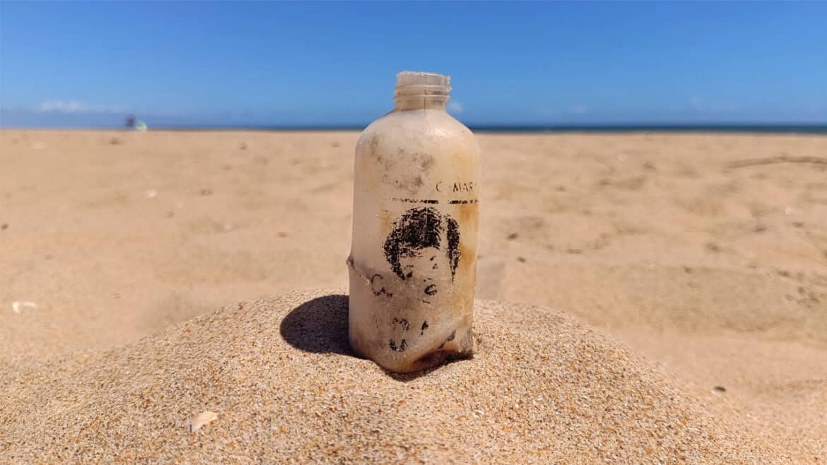 Una botella de plástico desgastada, con una etiqueta descolorida de «Borrador automático», se yergue en la arena de una playa desierta bajo un cielo azul y despejado.