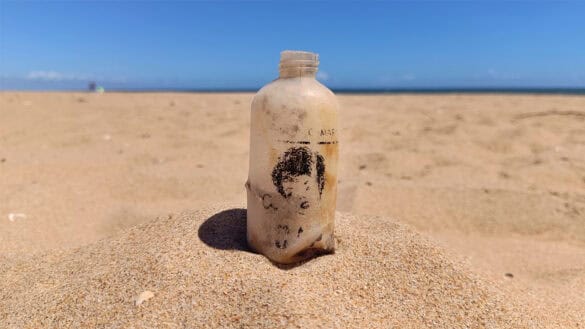Una botella de plástico desgastada, con una etiqueta descolorida de «Borrador automático», se yergue en la arena de una playa desierta bajo un cielo azul y despejado.