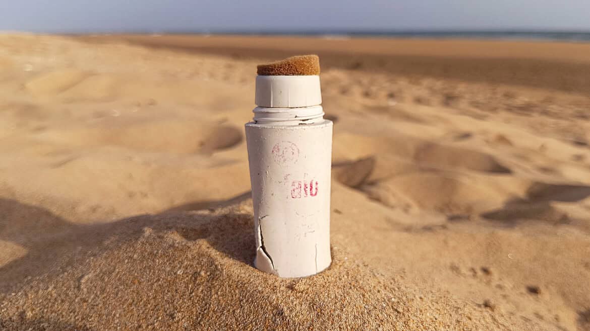 Un tubo blanco y gastado con una punta de esponja —que se asemeja a un borrador automático— se yergue en posición vertical, parcialmente enterrado en la arena de una playa, con el mar y el cielo como telón de fondo.