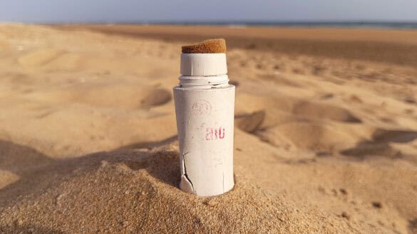 Un tubo blanco y gastado con una punta de esponja —que se asemeja a un borrador automático— se yergue en posición vertical, parcialmente enterrado en la arena de una playa, con el mar y el cielo como telón de fondo.