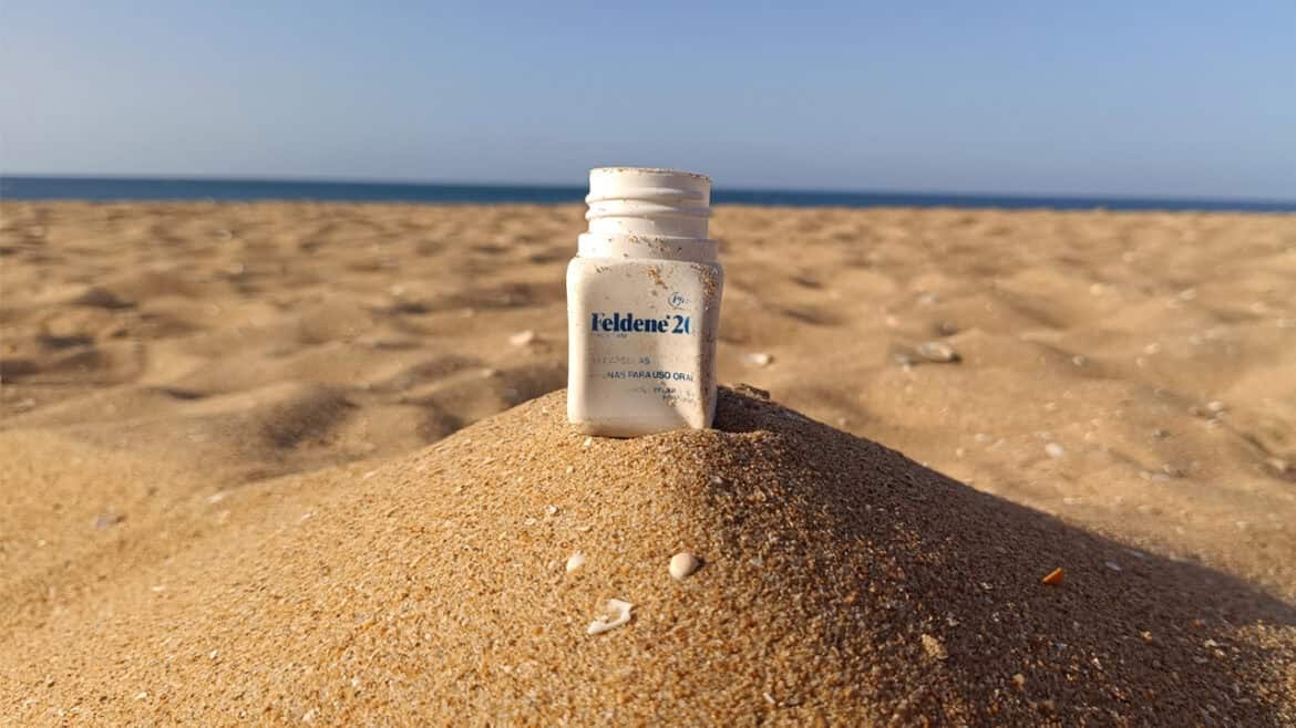 Un pequeño frasco blanco de Feldene al 2 %, que recuerda a los años 80, está parcialmente enterrado en posición vertical en un montículo de arena en una playa, con el mar y el cielo al fondo.