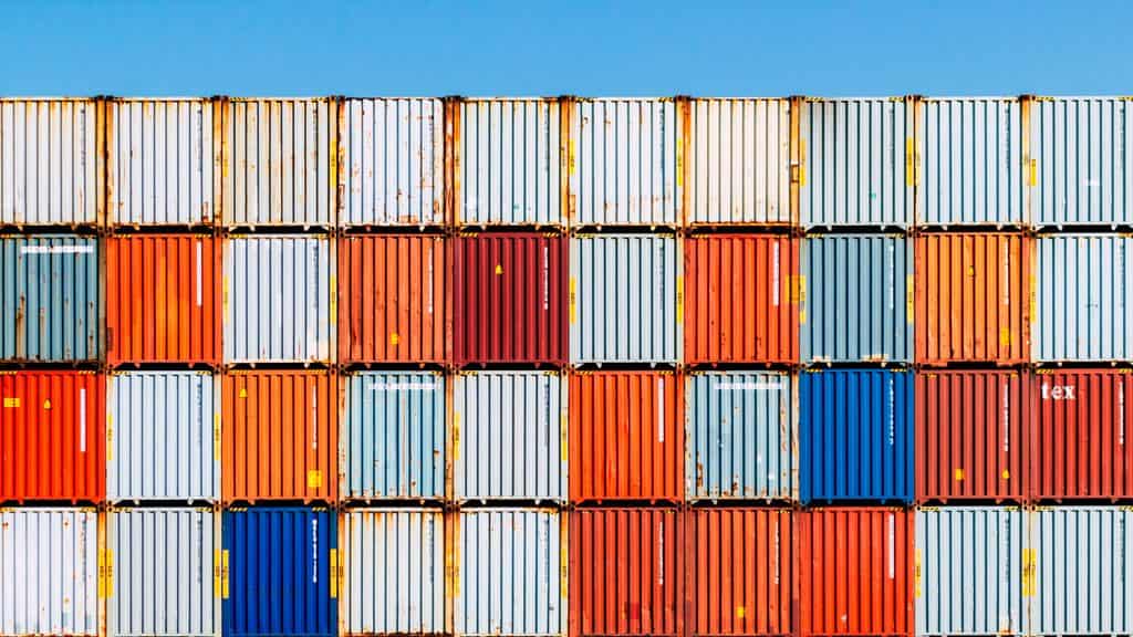 Contenedores de carga apilados de diversos colores se alinean en filas bajo un cielo azul claro, lo que pone de relieve un aspecto crucial del «viaje de los residuos», en el que el comercio mundial se entrecruza con problemas como la contaminación plástica.