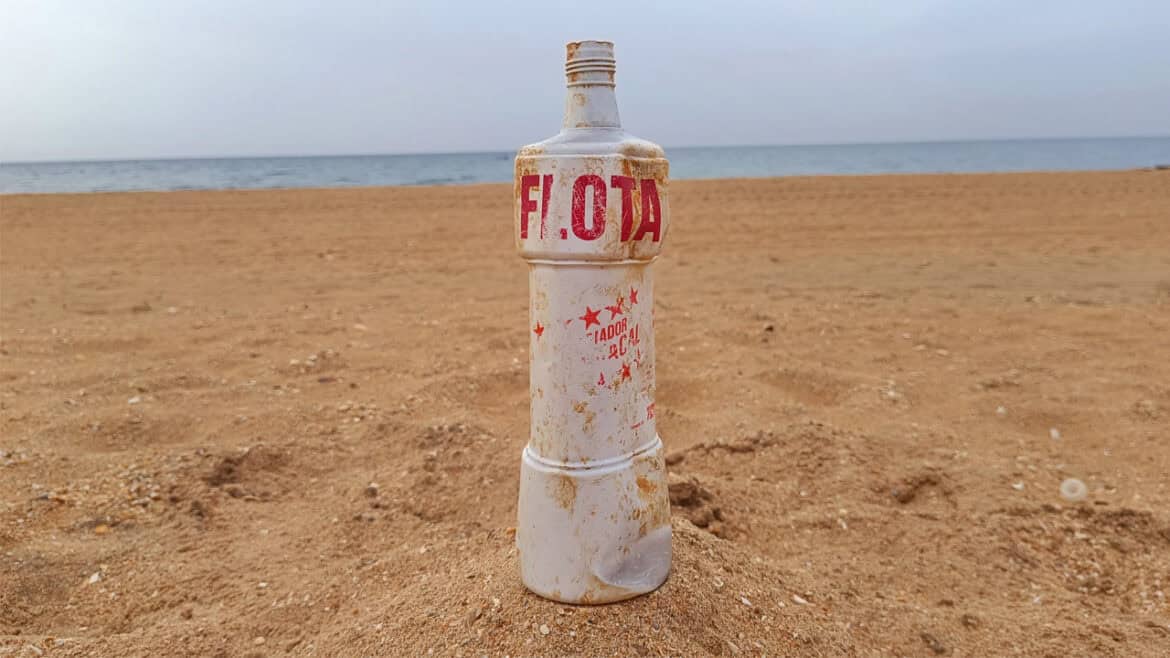 Una botella de plástico blanca de «Limpiador Flota», parcialmente enterrada en la arena de una playa, con letras rojas descoloridas y manchas de suciedad, evoca recuerdos de los años 80 con el océano y el cielo como telón de fondo.