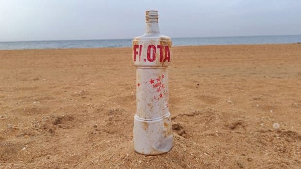 Una botella de plástico blanca de «Limpiador Flota», parcialmente enterrada en la arena de una playa, con letras rojas descoloridas y manchas de suciedad, evoca recuerdos de los años 80 con el océano y el cielo como telón de fondo.