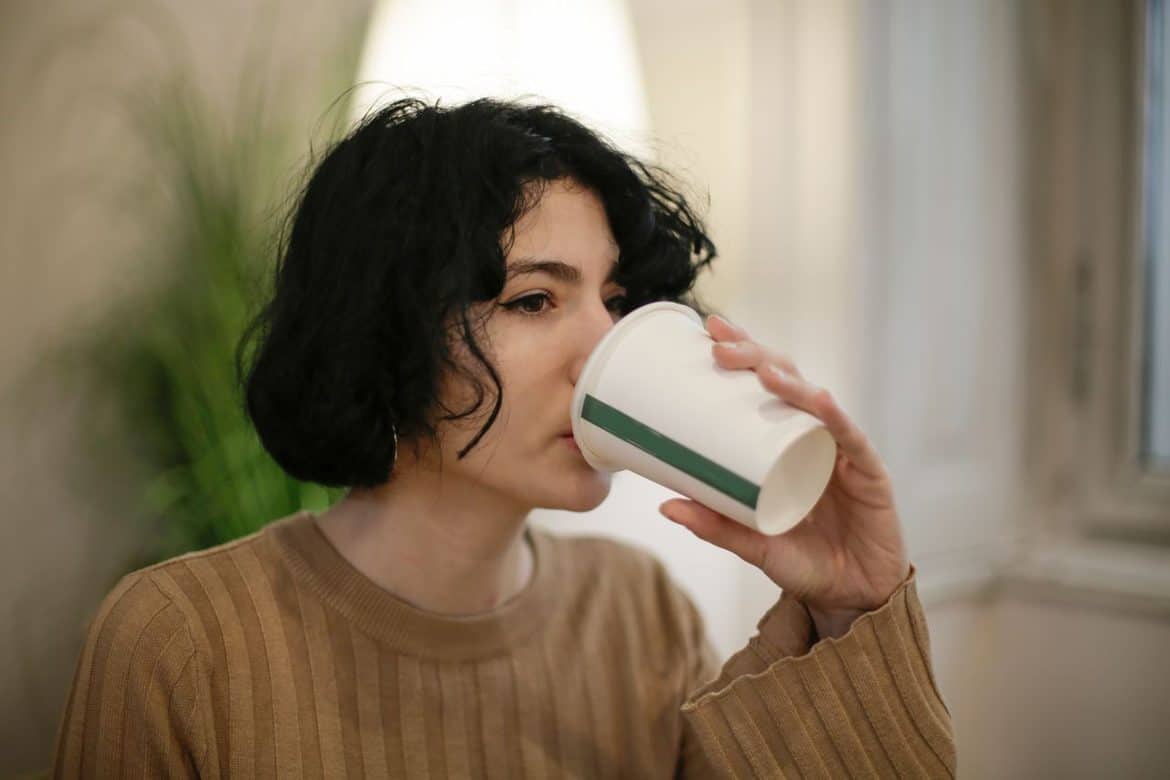 Una persona con el pelo corto y negro, que lleva un jersey marrón, bebe de un vaso de papel en el interior de una habitación, con una planta y una ventana al fondo.