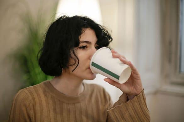 Una persona con el pelo corto y negro, que lleva un jersey marrón, bebe de un vaso de papel en el interior de una habitación, con una planta y una ventana al fondo.
